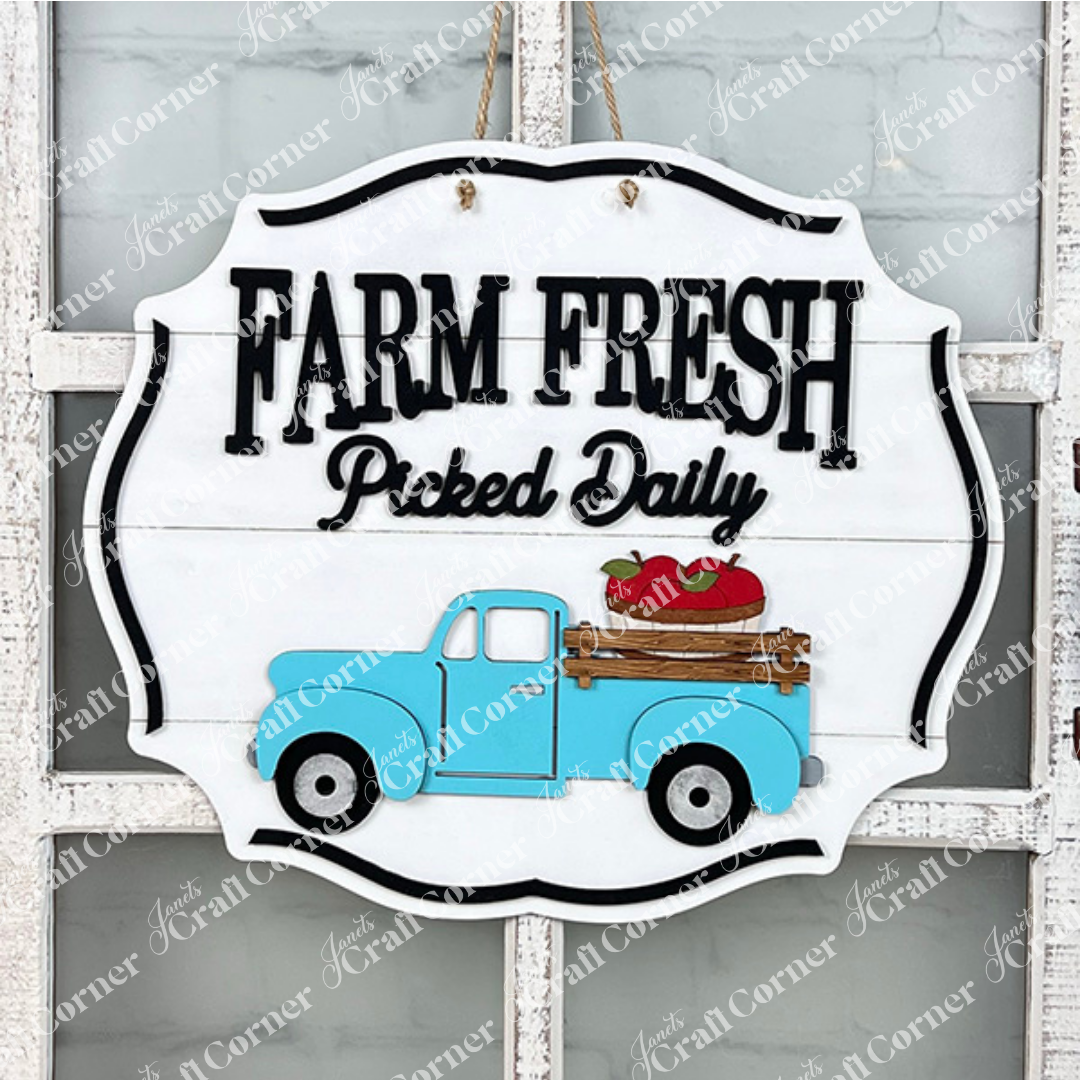 A rustic Farm Fresh Apples Sign by Janet's Craft Corner, featuring the bold black phrase "Farm Fresh Picked Daily" alongside a charming illustration of a blue vintage truck carrying red apples, is beautifully set against a white background.