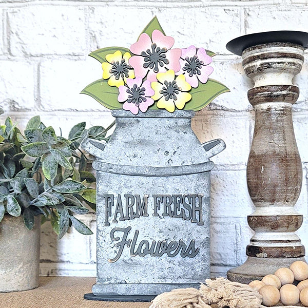 A galvanized milk jug featuring Janet's Craft Corner's "Farm Fresh Flowers Shelf Sitter" elegantly displayed, filled with pastel blooms and lush greenery, is positioned next to a wooden candle holder and a small plant against a white brick wall, radiating farmhouse charm.