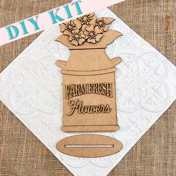 The image features the Farm Fresh Flowers Shelf Sitter from Janet's Craft Corner, a DIY kit that includes a wooden cutout of a galvanized milk jug overflowing with flowers. The phrase "Farm Fresh Flowers" is elegantly engraved on it. Set against textured burlap and decorative white tile, the design exudes farmhouse charm. "DIY Kit" is prominently displayed in the top left corner.