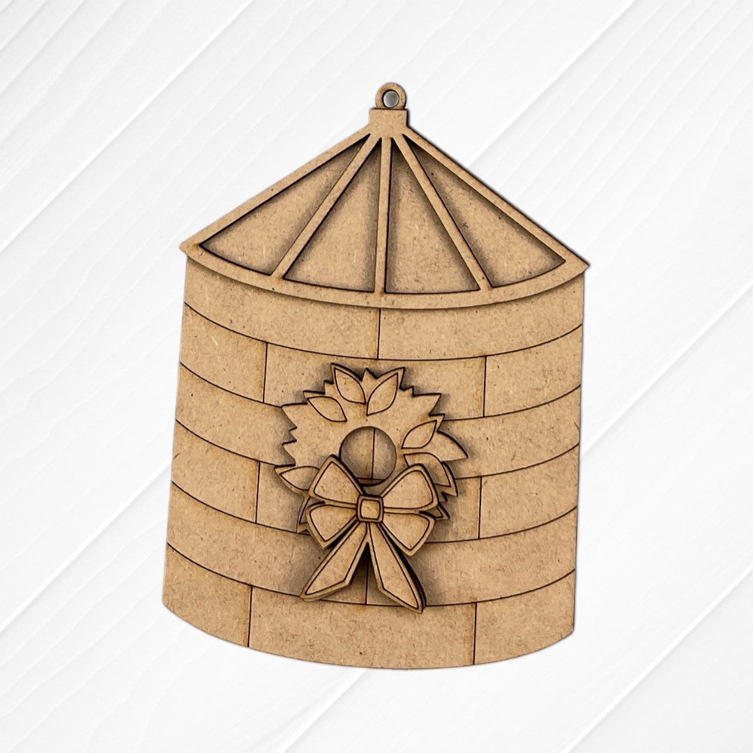 A wooden grain silo ornament with a pointed roof and wreath detail from Janet's Craft Corner—part of the Farm Life Christmas Ornaments DIY seasonal craft kit (choice of 4)—shown on a white wood-textured background.