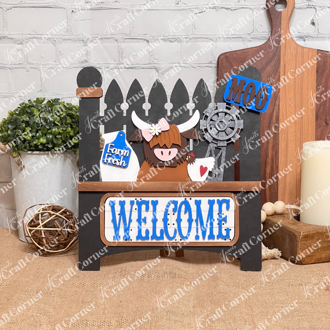 Janet's Craft Corner offers a DIY home decor kit: the Farm Welcome Interchangeable Insert, featuring a black garden fence base with "MOO" and "WELCOME" in blue, depicting a cartoon highland cow, milk bottle labeled "Farm Fresh," rusty windmill, and heart cutout. Perfect for burlap surfaces against white brick walls.