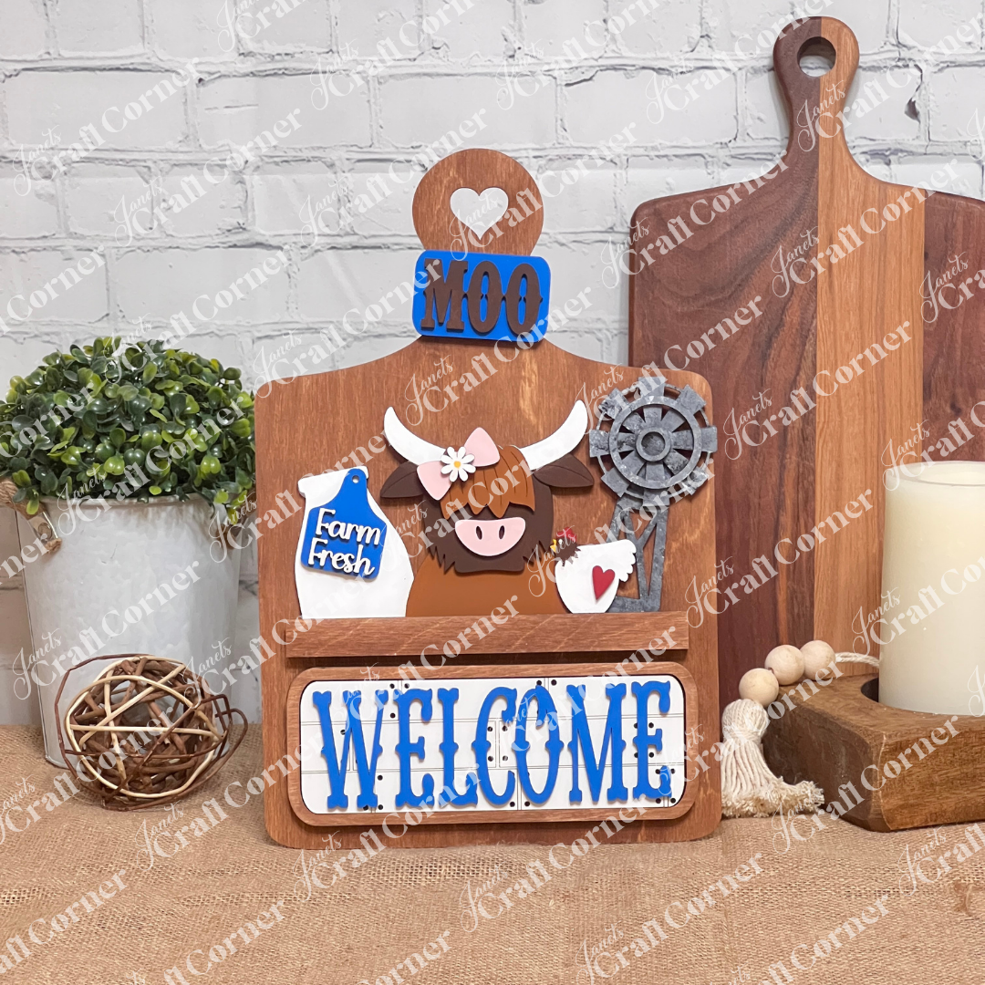 Janet's Craft Corner offers a DIY home decor kit: the Farm Welcome Interchangeable Insert, featuring a wooden board with "MOO" and "WELCOME" in blue, depicting a cartoon highland cow, milk bottle labeled "Farm Fresh," rusty windmill, and heart cutout. Perfect for burlap surfaces against white brick walls.