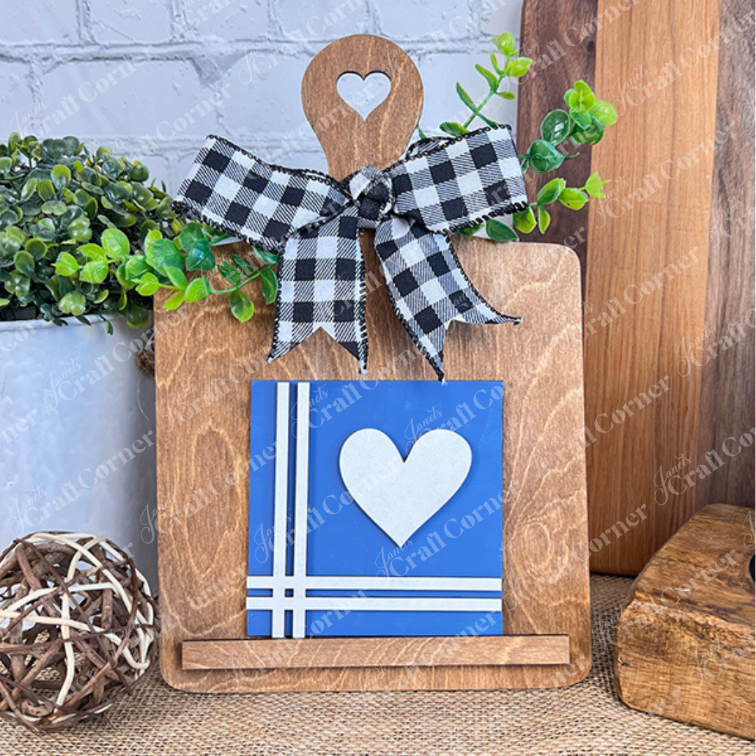 The Home Squares by Janet's Craft Corner features a wooden cutting board adorned with a black-and-white checkered bow, faux greenery, and a blue Swappable Square with white stripes and heart—perfect for rustic tiered tray decor and charming wood accents.