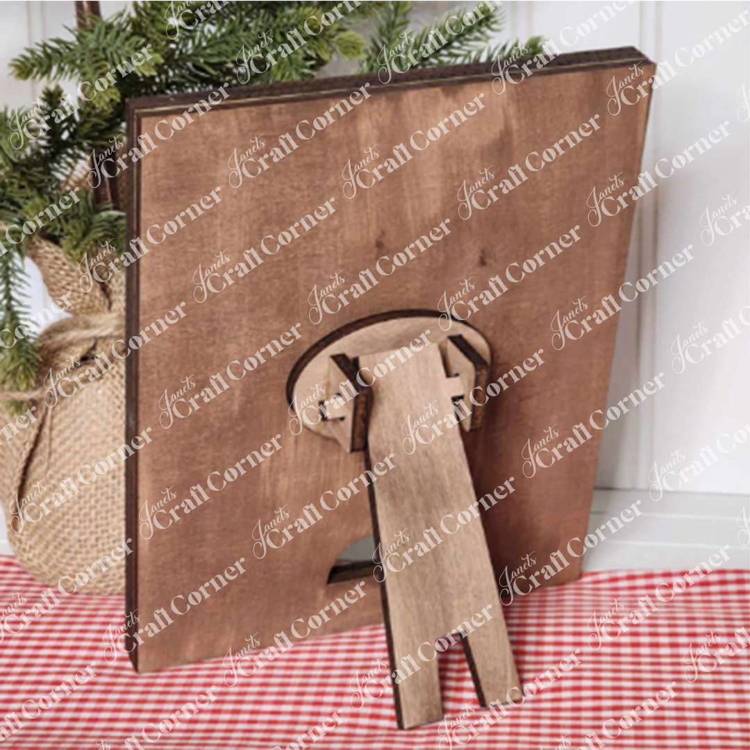 A Farmhouse Frame for Interchangeable Signs by Janet's Craft Corner is elegantly displayed on a checkered red and white cloth, capturing the essence of farmhouse decor. A small plant in a burlap sack adds to its rustic charm. This frame is ideal for featuring your latest DIY home decor craft kit creations or swapping out signs to match any season.