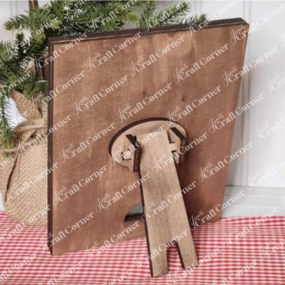 A Farmhouse Frame for Interchangeable Signs by Janet's Craft Corner is elegantly displayed on a checkered red and white cloth, capturing the essence of farmhouse decor. A small plant in a burlap sack adds to its rustic charm. This frame is ideal for featuring your latest DIY home decor craft kit creations or swapping out signs to match any season.