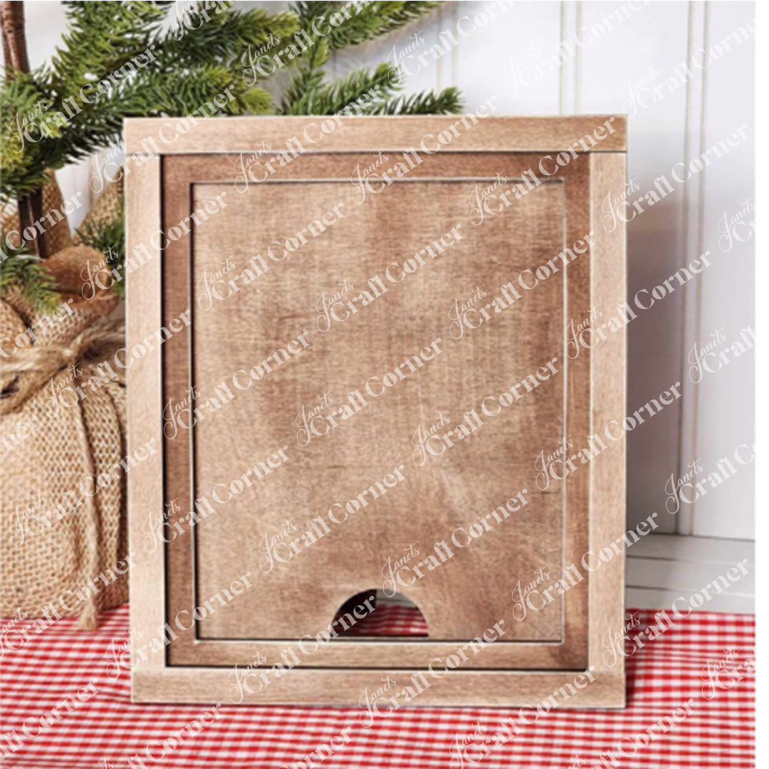A DIY home decor craft kit featuring the Farmhouse Frame for Interchangeable Signs from Janet's Craft Corner stands upright on a checkered red and white cloth. In the background, there's a potted evergreen tree wrapped in burlap, adding a touch of farmhouse decor to the scene.