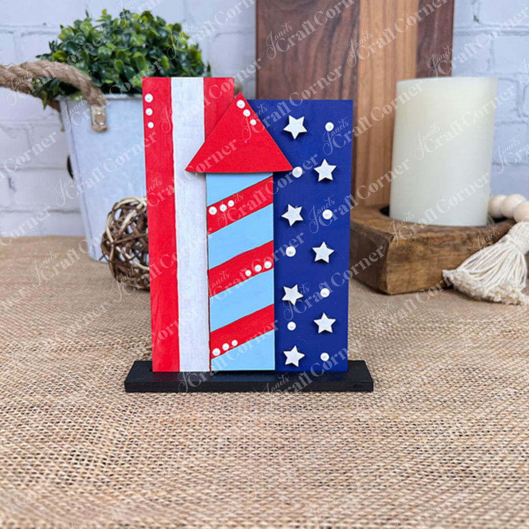 The Patriotic Mini Shelf Sitters – DIY Craft Kit from Janet's Craft Corner features firework-shaped wooden pieces in Americana style, painted with red and white stripes, blue stars, and a black base—perfect patriotic decor for your home.