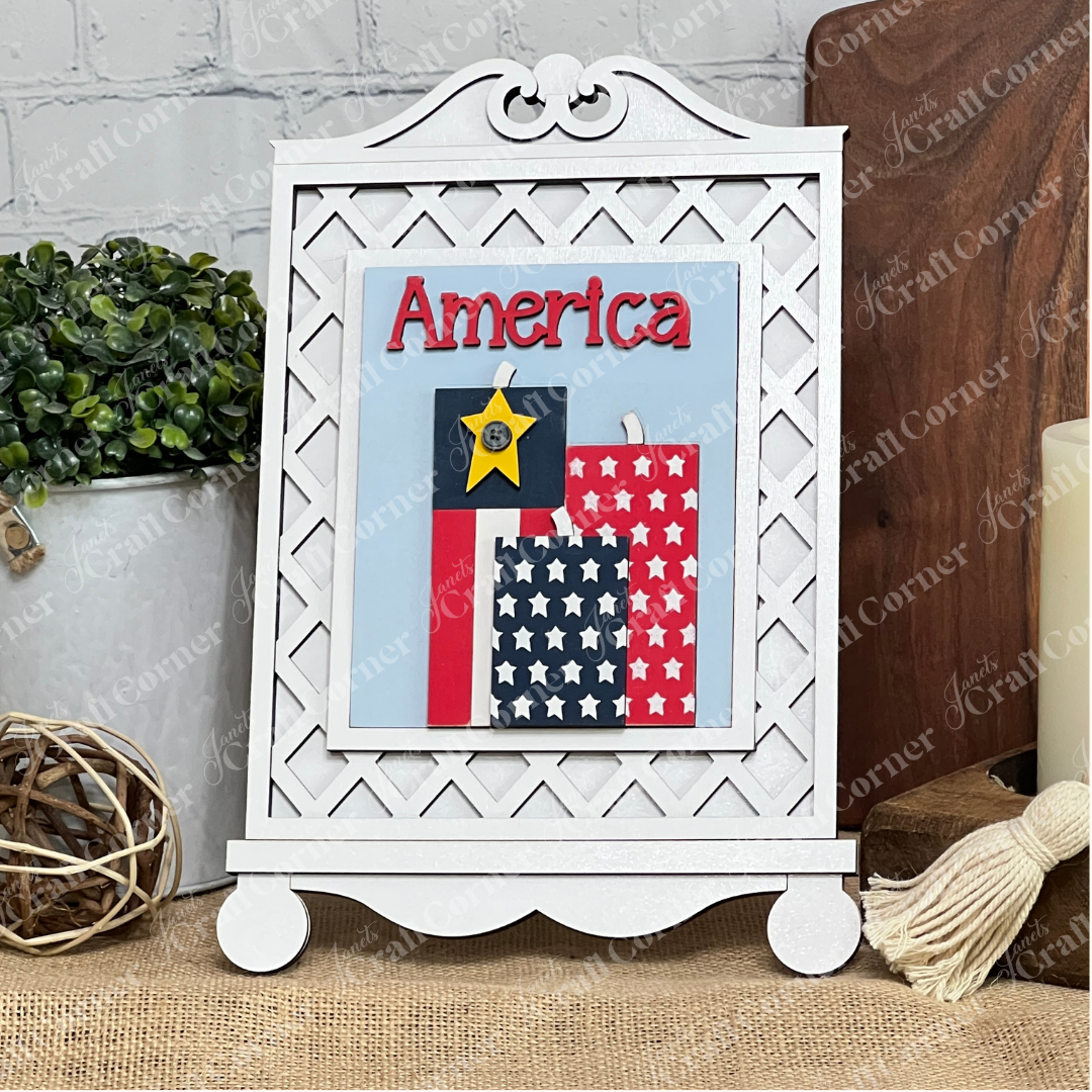 Janet's Craft Corner Patriotic Farmhouse Signs DIY kit features a framed “America” with three red, white, blue, and black tag accents and stars—perfect for tiered tray or spring farmhouse decor—displayed on a white lattice easel.
