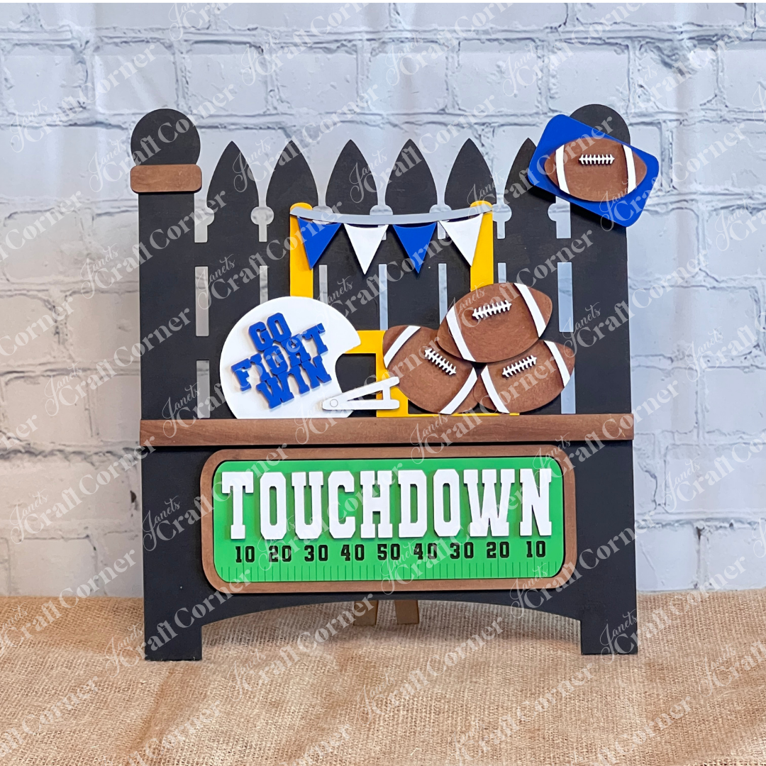 Janet's Craft Corner offers a Garden Fence with Insert DIY kit featuring a football-themed display. It includes a picket fence with pennants, footballs, and a "Go Fight Win" sign above a burlap surface. A green scoreboard-style sign reads "TOUCHDOWN," all set against a rustic brick wall backdrop.