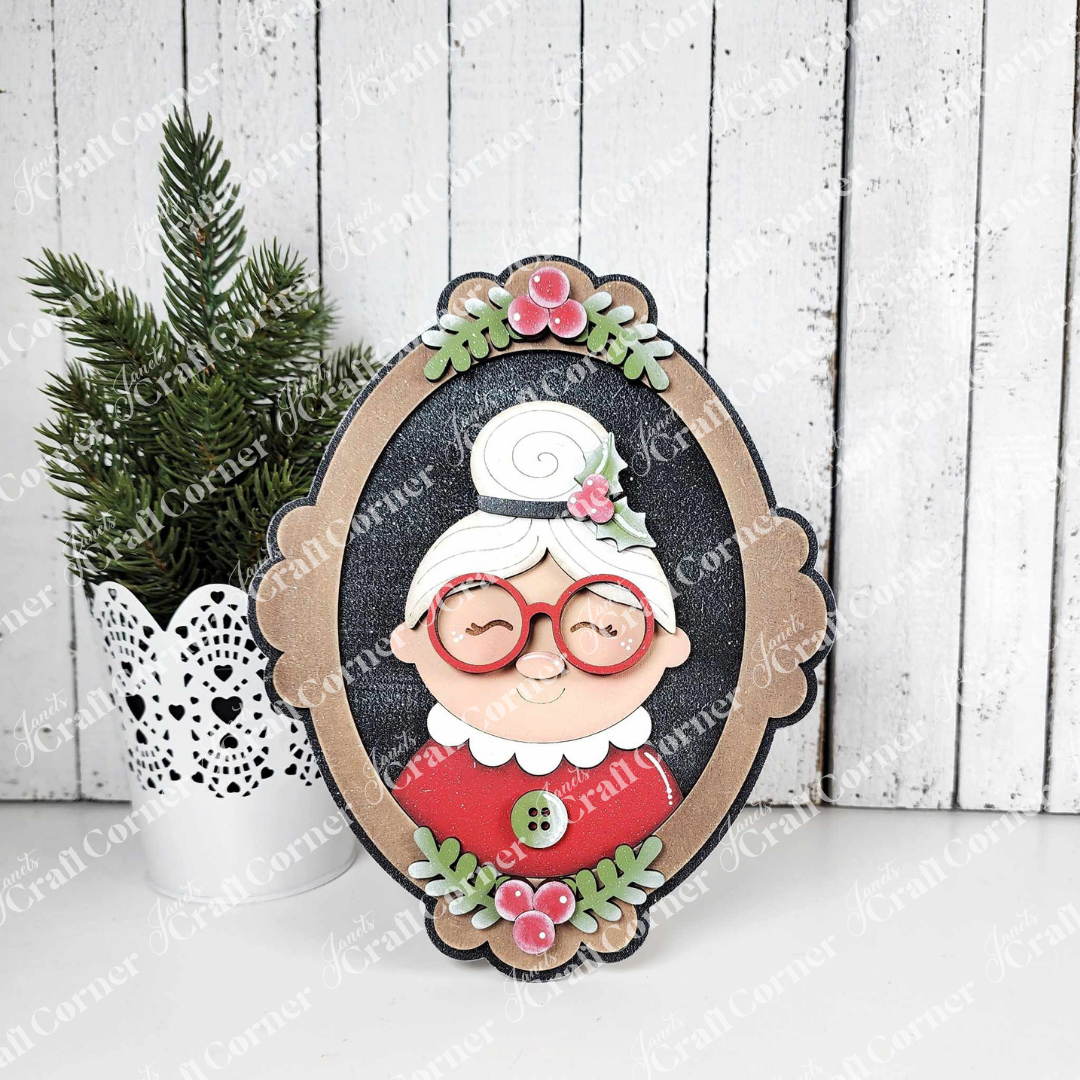 The Framed Mrs. Claus Shelf Sitter by Janet's Craft Corner features a cartoon-style portrait of a smiling elderly woman with white hair, red glasses, and a pink flower—ideal as festive home decor or a charming shelf accent.
