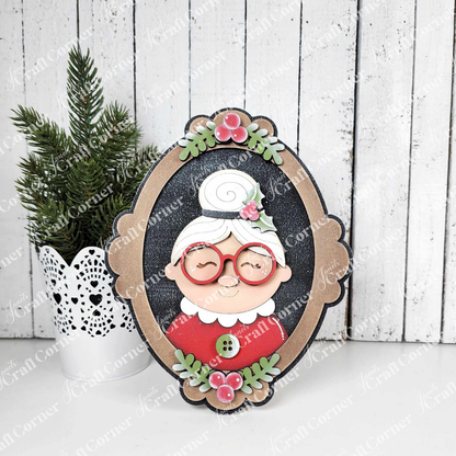 The Framed Mrs. Claus Shelf Sitter by Janet's Craft Corner features a cartoon-style portrait of a smiling elderly woman with white hair, red glasses, and a pink flower—ideal as festive home decor or a charming shelf accent.
