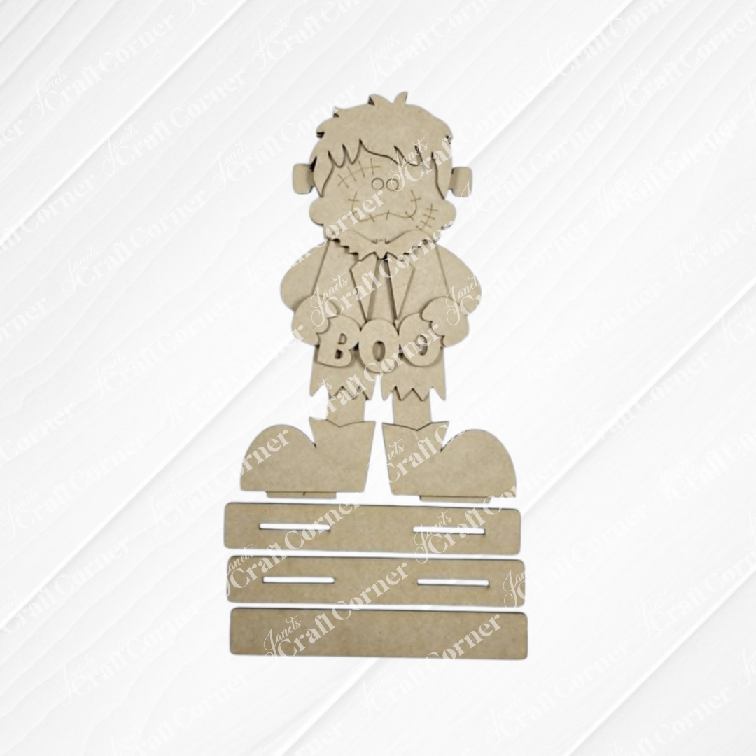 The Frankie Shelf Sitter DIY home decor craft kit from Janet’s Craft Corner features a cartoon Frankenstein cutout holding a “BOO” sign and standing on stacked rectangular bases, all made from plain brown cardboard—a delightful set of 1 that’s perfect for creating handmade Halloween decorations for your home.