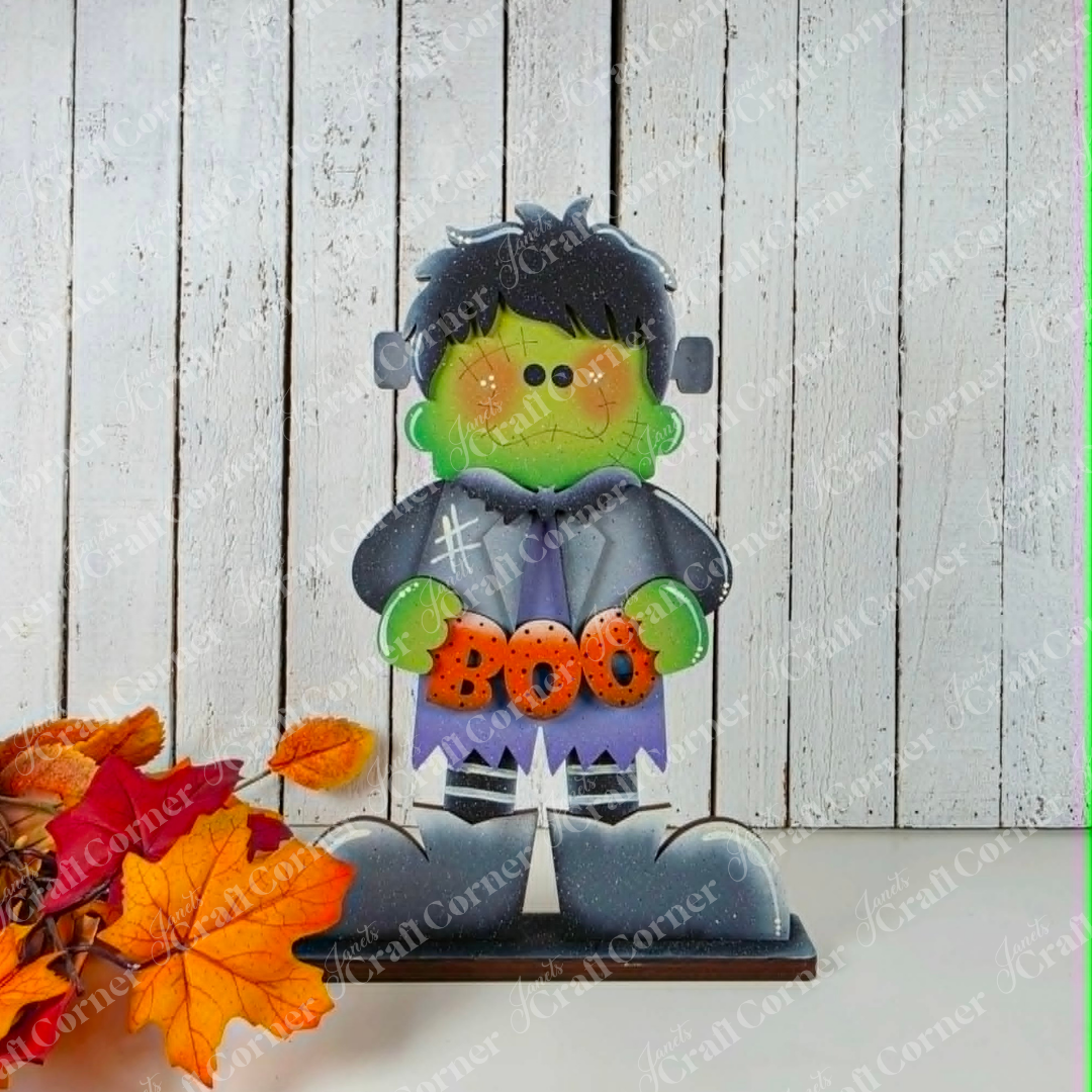 Add a playful touch to your Halloween decor with the Frankie Shelf Sitter from Janet’s Craft Corner—a DIY home decor craft kit that features a charming cartoon Frankenstein holding a “BOO” sign. This delightful figure stands on a wooden surface surrounded by autumn leaves against a white wooden plank background, making it perfect for displaying as part of your craft kit collection.