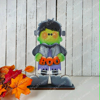 Add a playful touch to your Halloween decor with the Frankie Shelf Sitter from Janet’s Craft Corner—a DIY home decor craft kit that features a charming cartoon Frankenstein holding a “BOO” sign. This delightful figure stands on a wooden surface surrounded by autumn leaves against a white wooden plank background, making it perfect for displaying as part of your craft kit collection.