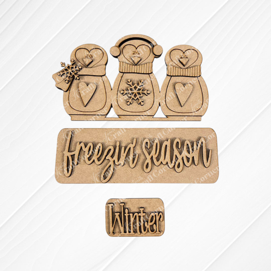 Janet's Craft Corner presents the "Freezin' Season" Interchangeable Insert, a DIY home decor kit featuring wooden cutouts on a textured background with charming penguins wearing winter accessories and decorated with heart-shaped designs. The script "Freezin Season" appears alongside a plaque labeled "Winter," making it an ideal craft kit for versatile home decoration.