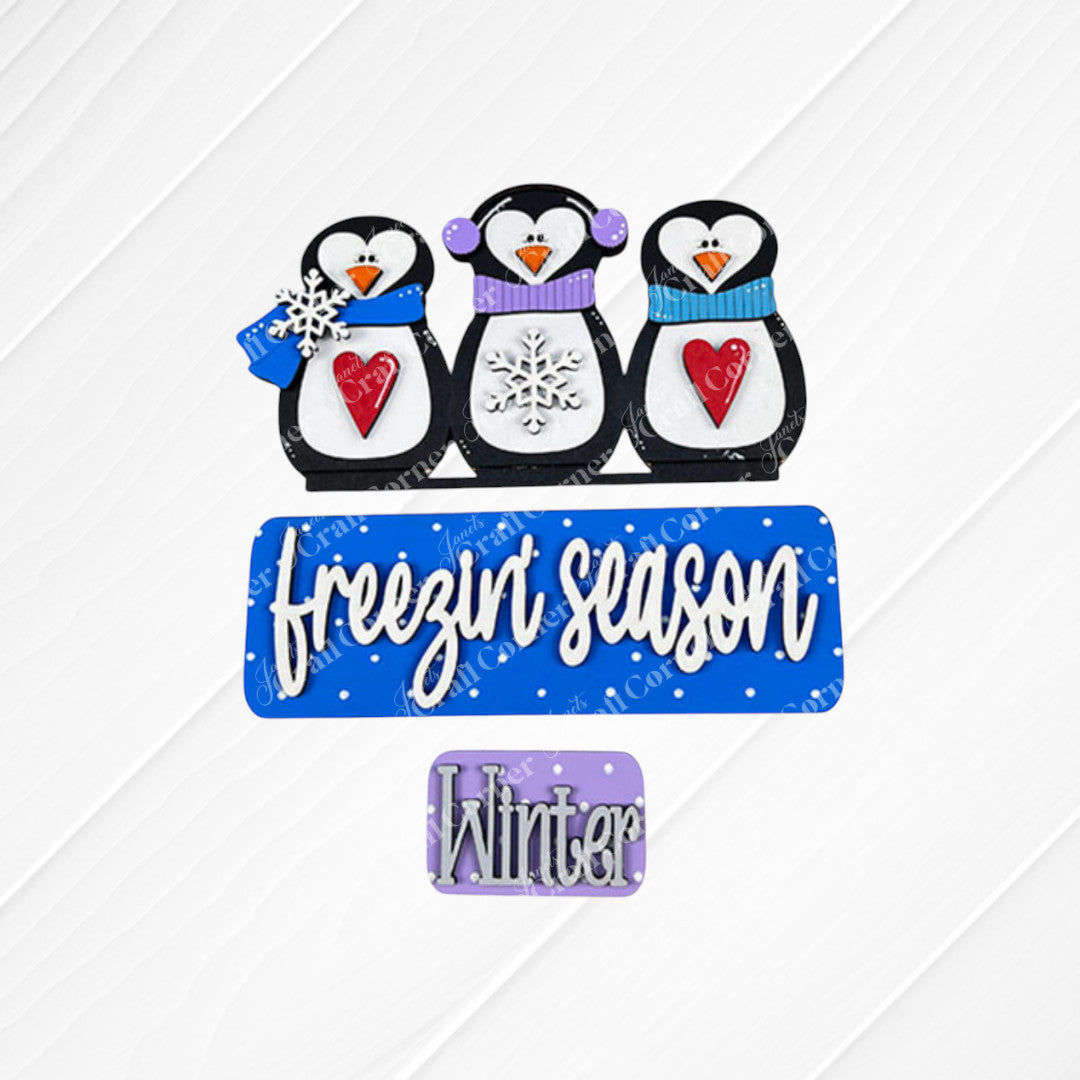 The Freezin' Season Interchangeable Insert by Janet's Craft Corner features three playful penguin figures dressed in charming hats and scarves, each holding colorful hearts and snowflakes. The decorative square showcases a blue sign with the text "freezin' season" above another purple sign labeled "Winter." This delightful DIY home decor kit is perfect for adding versatility to your home styling.