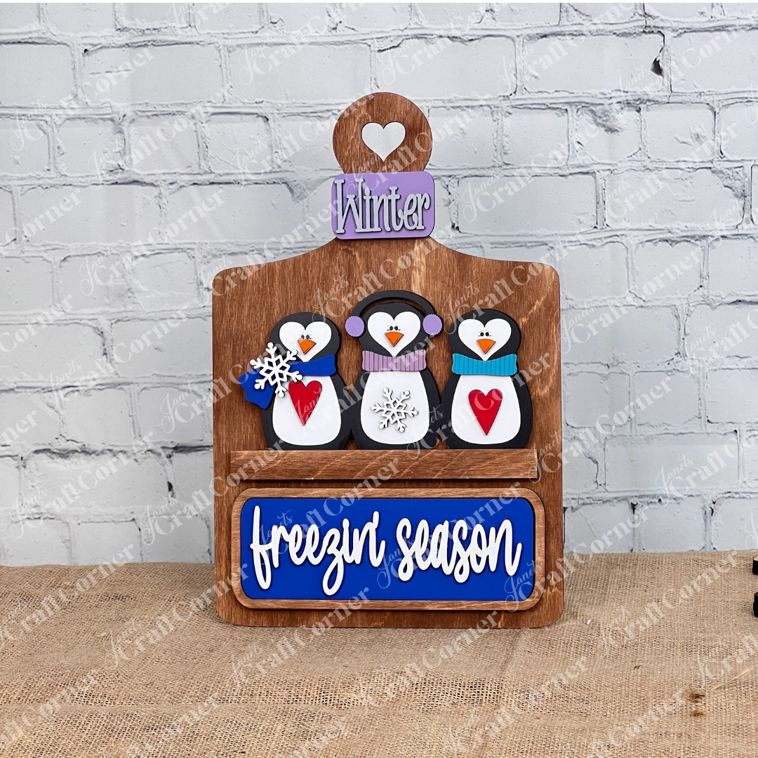 Janet's Craft Corner offers the "Freezin' Season" interchangeable insert, a 3-piece DIY seasonal decor kit featuring hand-painted penguins in earmuffs and scarves, a "Winter" sign on a brown base, and "freezin' season" text on blue below. Ideal for home decor enthusiasts!.