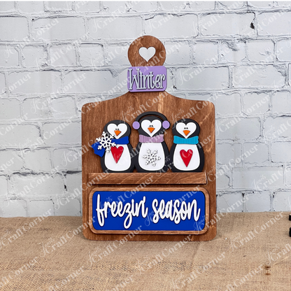 Janet's Craft Corner offers the "Freezin' Season" interchangeable insert, a 3-piece DIY seasonal decor kit featuring hand-painted penguins in earmuffs and scarves, a "Winter" sign on a brown base, and "freezin' season" text on blue below. Ideal for home decor enthusiasts!.