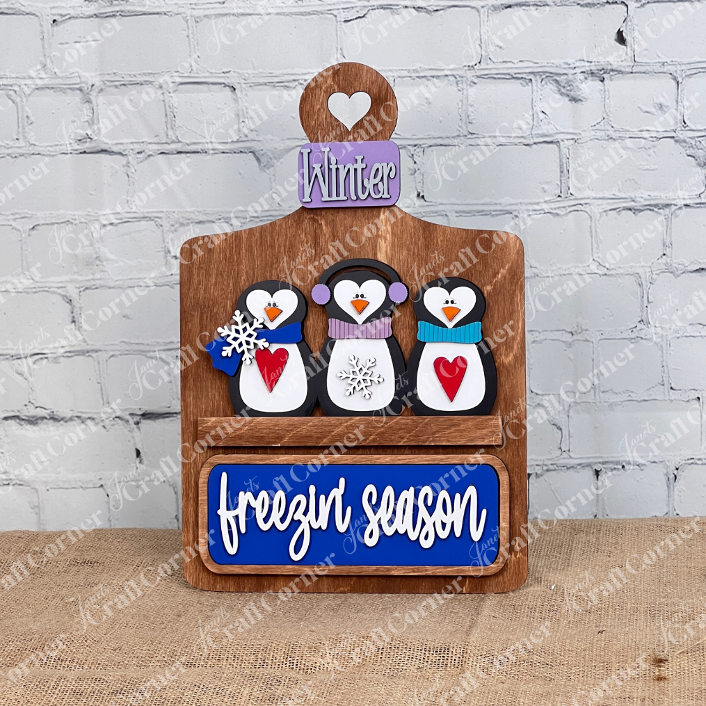 A charming DIY home decor project from Janet's Craft Corner, this wooden breadboard base insert features three penguins with earmuffs and scarves, each holding a red heart. The middle penguin has a snowflake, topped by a "Winter" sign and "freezin' season" on a blue plaque, set against a white brick wall.