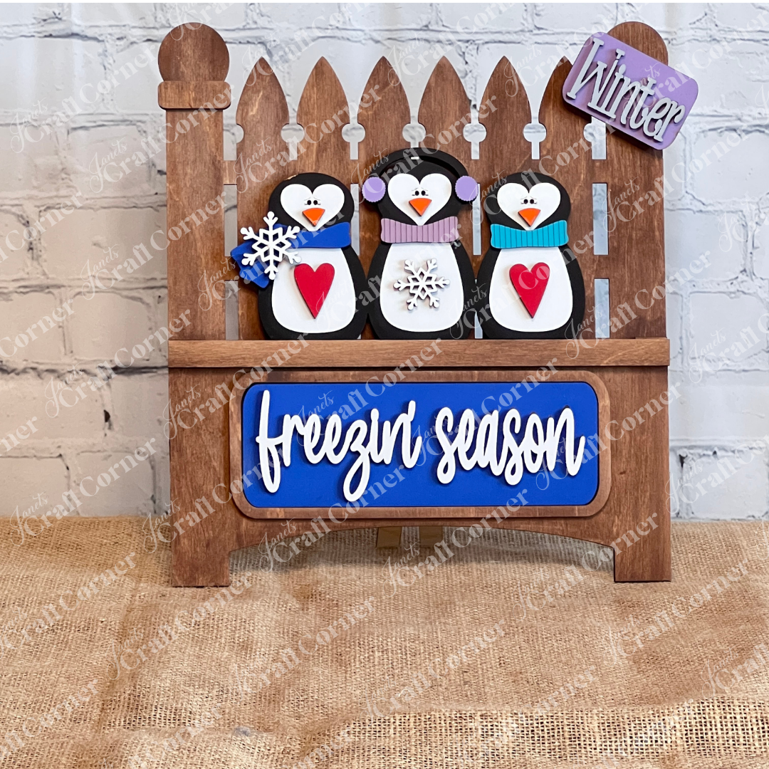 Janet's Craft Corner offers the "Freezin' Season Interchangeable Insert," a 3-piece DIY craft kit featuring a wooden decoration with hand-painted penguins in winter gear and red hearts on a blue plaque. A small "Winter" sign hangs above, perfect for interchangeable home decor enthusiasts.