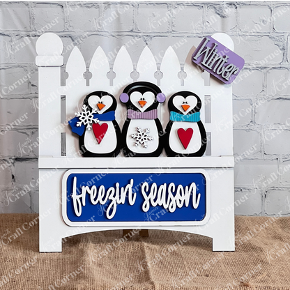 The Freezin' Season Interchangeable Insert by Janet's Craft Corner is a DIY seasonal home decor craft kit, featuring three hand-painted penguins on a white fence backdrop with scarves and heart details. A "freezin' season" and "Winter" sign complete this charming burlap display.