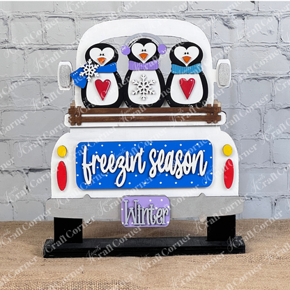 Janet's Craft Corner offers a DIY interchangeable home decor kit featuring three penguins in scarves on an antique truck. With "freezin' season" and "Winter" on the license plate, this charming piece, set against a white brick wall, adds a cozy touch to any space.