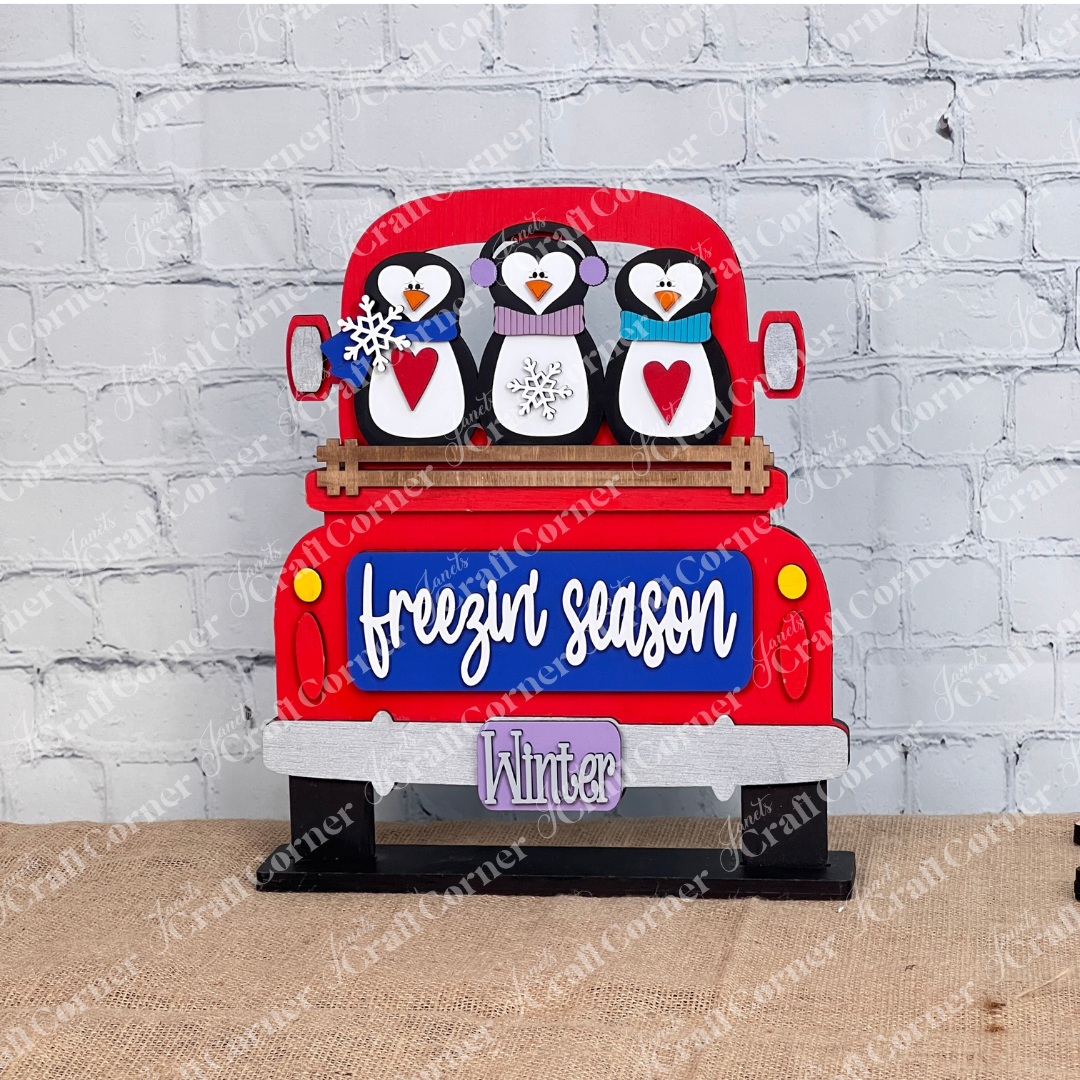 Janet's Craft Corner offers the Freezin' Season Interchangeable Insert, a DIY seasonal home decor craft kit featuring a hand-painted red truck with penguins in winter gear and "freezin’ season" on display. Ideal for interchangeable home decor enthusiasts, set against a white brick wall.