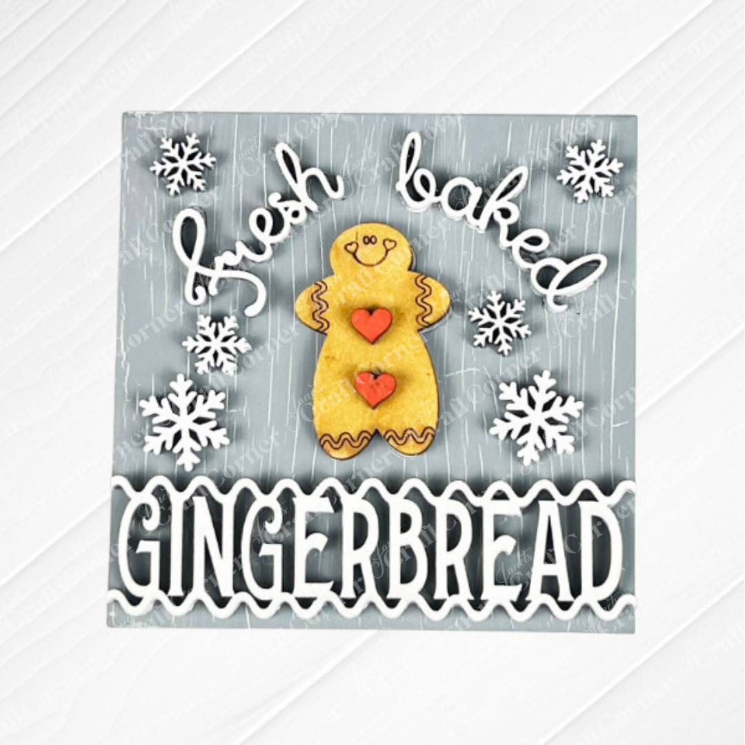 Janet's Craft Corner Gingerbread Squares feature a smiling gingerbread cookie, red heart buttons, and white snowflakes on gray wood with "fresh baked GINGERBREAD" in playful white text. Perfect DIY seasonal decor or for Swappable Square Displays.