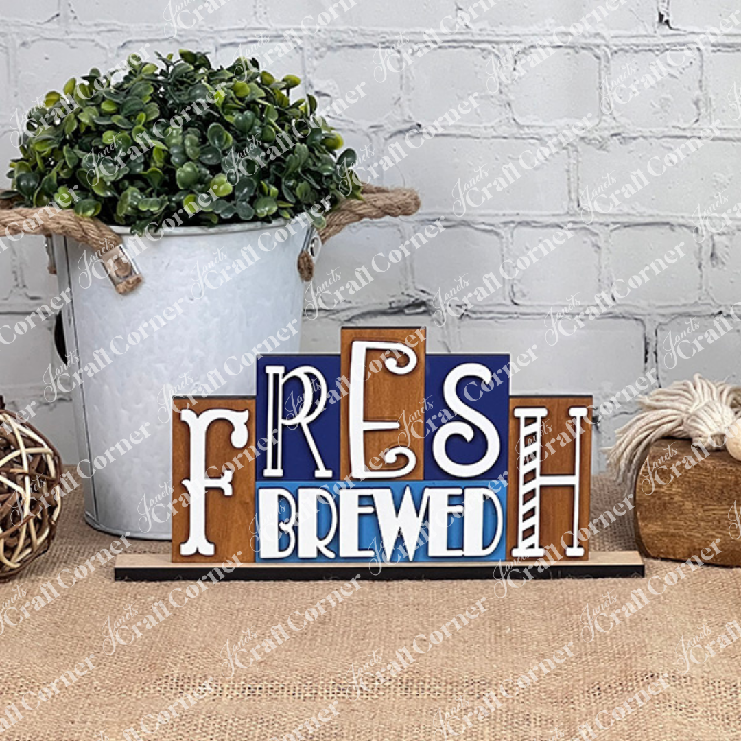 Introducing the Fresh Brewed Mini Word Block from Janet's Craft Corner, a delightful home décor piece. This DIY kit showcases a decorative sign featuring the words "Fresh Brewed" in an assortment of fonts and colors set against a wooden backdrop. It is elegantly displayed on a burlap-covered surface with a chic white brick wall providing the perfect background.