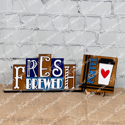 Introducing the Fresh Brewed Mini Word Block from Janet's Craft Corner, a delightful home décor piece. This DIY kit showcases a decorative sign featuring the words "Fresh Brewed" in an assortment of fonts and colors set against a wooden backdrop. It is elegantly displayed on a burlap-covered surface with a chic white brick wall providing the perfect background. included in the photo is also one of our Coffee Swappable Squares.