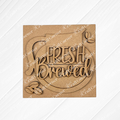 The Coffee Bar Swappable Squares DIY craft kit by Janet's Craft Corner features a wooden coffee pot cutout with "Fresh Brewed" and four coffee beans on a textured white background—perfect seasonal home decor for coffee lovers.