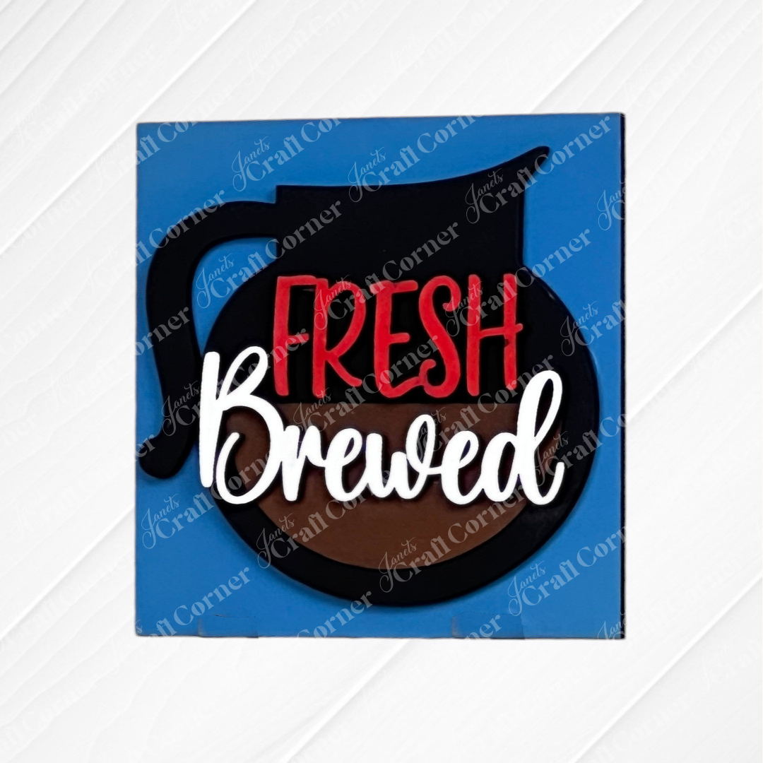 A blue square sign from Janet's Craft Corner's Coffee Bar Swappable Squares DIY seasonal home decor kit displays a coffee pot graphic and "Fresh Brewed" in red and white, on a white wooden surface.