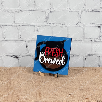 A blue square sign from Janet's Craft Corner's Coffee Bar Swappable Squares DIY seasonal home decor kit displays a coffee pot graphic and "Fresh Brewed" in red and white, on a wooden stand against a white brick wall and burlap surface.