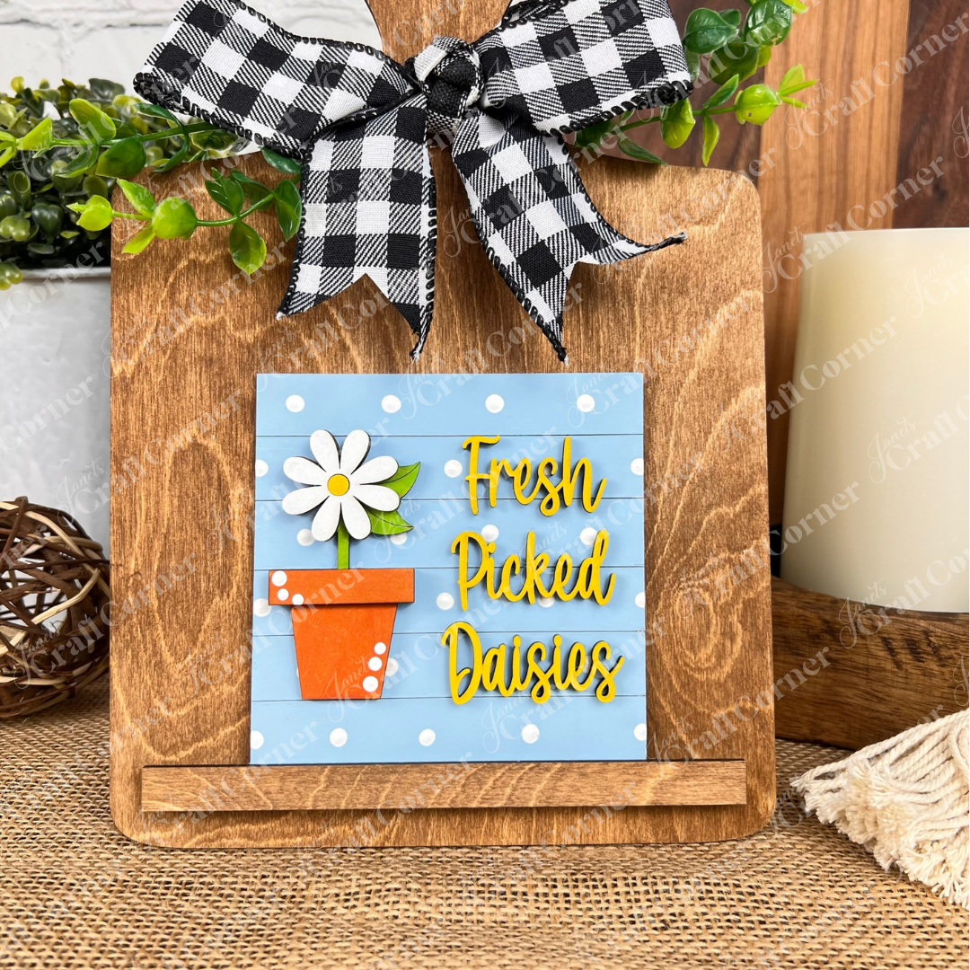 A wooden cutting board with a black-and-white checkered bow displays Janet's Craft Corner Bloom & Grow Squares, featuring a daisy in an orange pot and “Fresh Picked Daisies” on blue, accented with greenery for charming tiered tray decor.