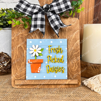 A wooden cutting board with a black-and-white checkered bow displays Janet's Craft Corner Bloom & Grow Squares, featuring a daisy in an orange pot and “Fresh Picked Daisies” on blue, accented with greenery for charming tiered tray decor.