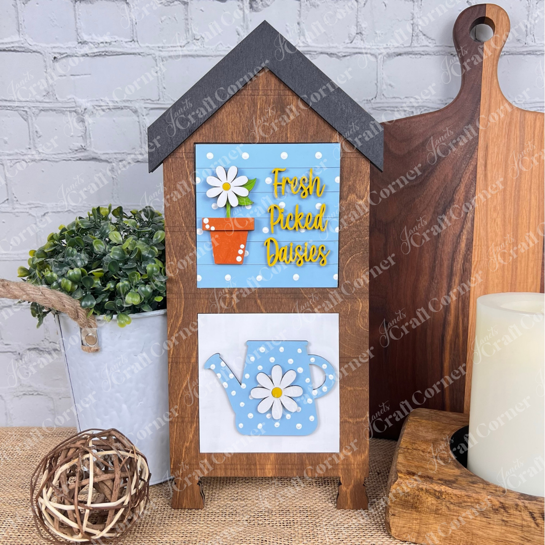 Janet's Craft Corner Bloom & Grow Squares: House-shaped wood sign kit featuring “Fresh Picked Daisies” in yellow above an orange flower pot with daisies and a blue polka-dot watering can—perfect for tiered tray decor.