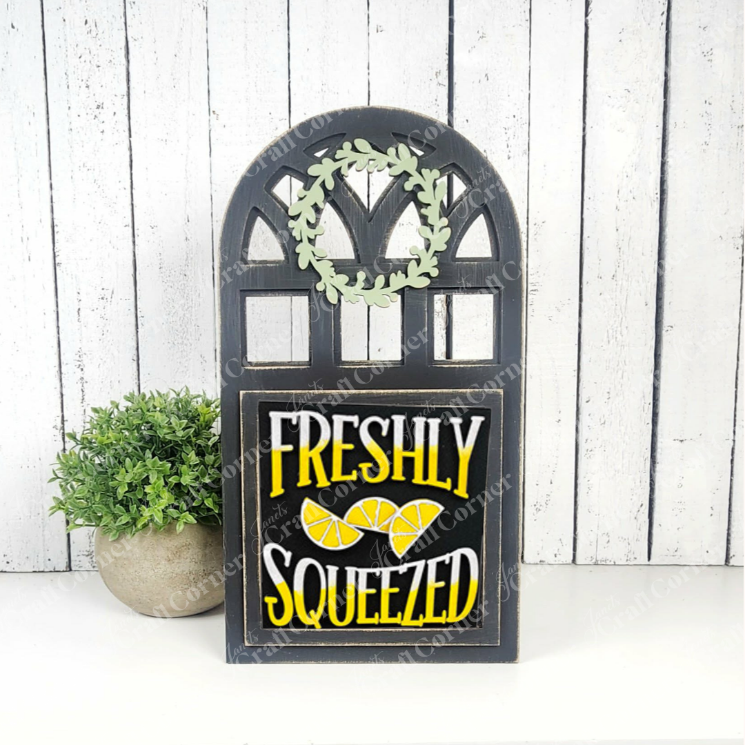 Janet's Craft Corner's Lemon Squares is a decorative black sign with "Freshly Squeezed" in bold white and yellow letters and lemon slices—perfect seasonal DIY decor for your home, shown against a white wooden wall with a green potted plant.