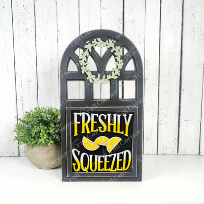 Janet's Craft Corner's Lemon Squares is a decorative black sign with "Freshly Squeezed" in bold white and yellow letters and lemon slices—perfect seasonal DIY decor for your home, shown against a white wooden wall with a green potted plant.