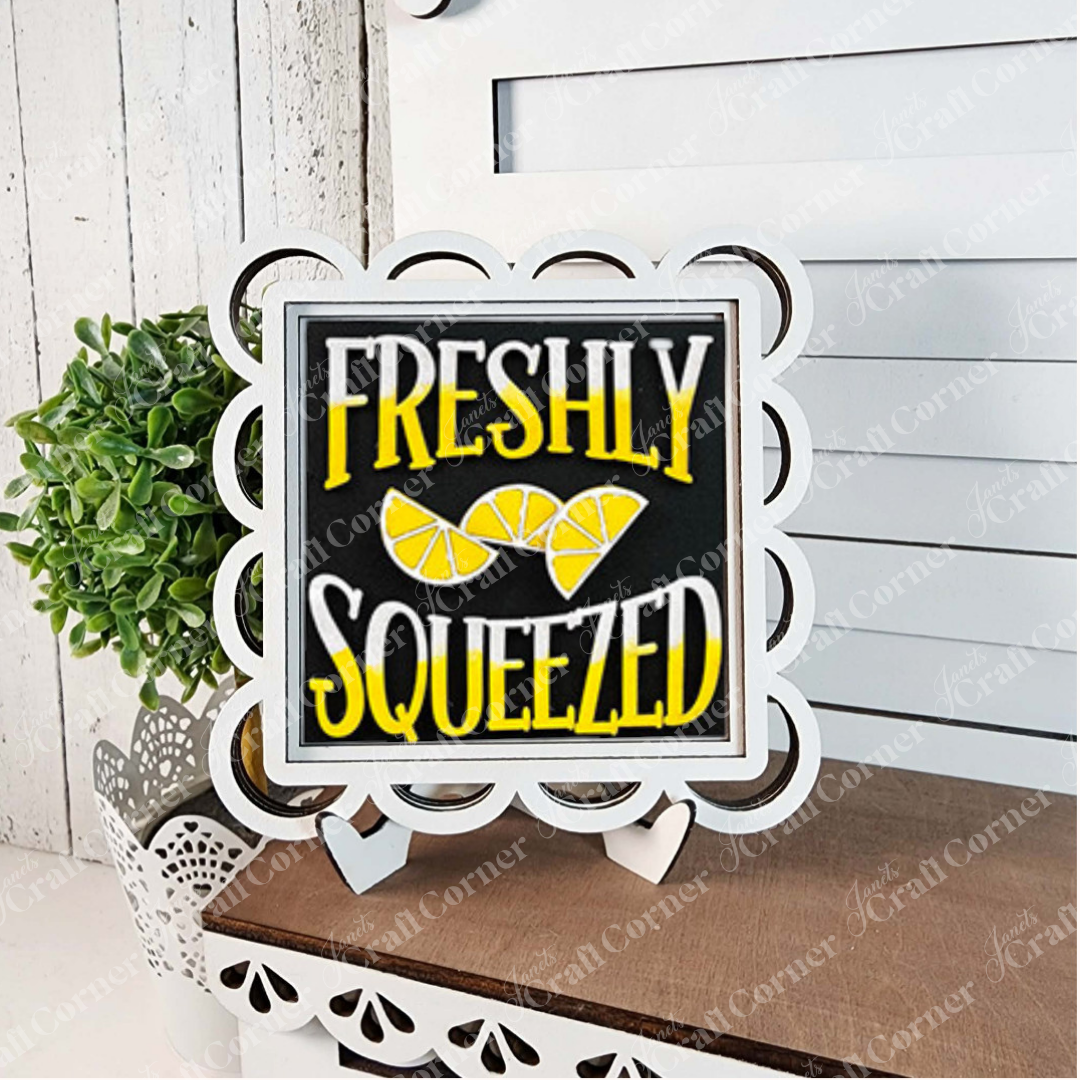 The "Lemon Squares" sign by Janet's Craft Corner features scalloped edges, yellow and white "Freshly Squeezed" text, and lemon slice art. Displayed on a white easel next to a potted plant, it’s perfect seasonal DIY home decor.