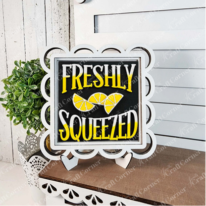 The "Lemon Squares" sign by Janet's Craft Corner features scalloped edges, yellow and white "Freshly Squeezed" text, and lemon slice art. Displayed on a white easel next to a potted plant, it’s perfect seasonal DIY home decor.