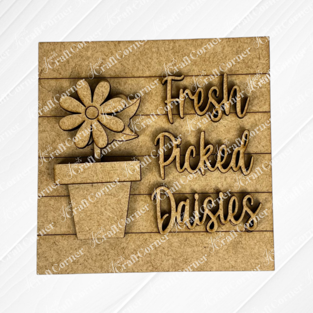 The Bloom & Grow Squares by Janet's Craft Corner is a layered wooden sign with a daisy in a pot and "Fresh Picked Daisies" in playful script, making it perfect for tiered tray decor on a slatted background.