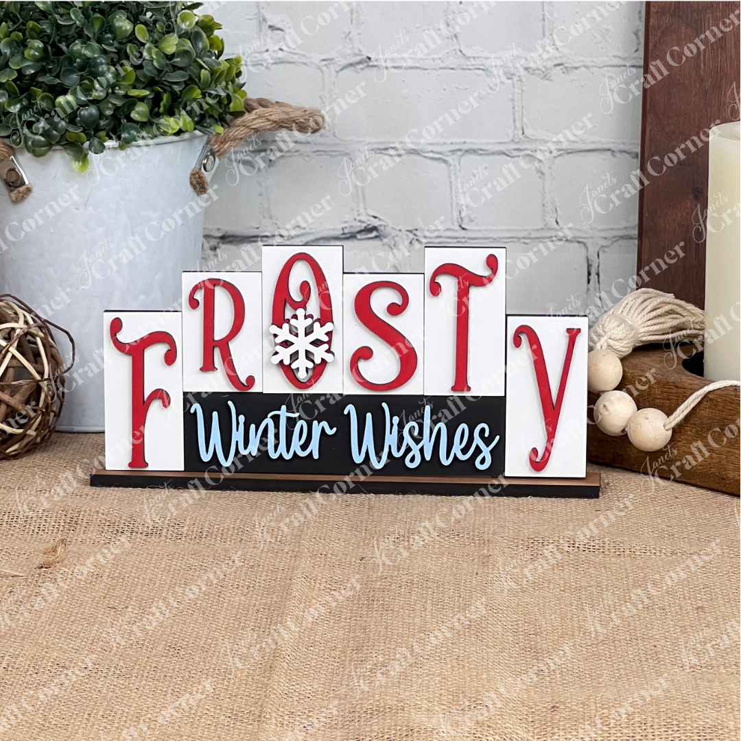 The Frosty Mini Word Block Shelf Sitter from Janet's Craft Corner is a delightful DIY decor kit that features "Frosty Winter Wishes" in red and white letters with a charming snowflake design. It brings a festive flair to your home decor when placed on a burlap-covered surface next to a white pot filled with greenery, accompanied by a woven ball and candle on a wooden tray against the backdrop of a brick wall.