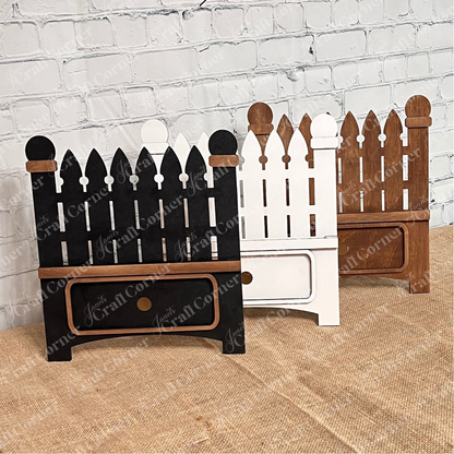 Three small picket fence-shaped decorations from Janet's Craft Corner rest on burlap against a white brick wall. These black, white, and brown DIY interchangeable bases each have a spots for the 3 pieces of a classic interchangeable insert, perfect for personalized home décor.