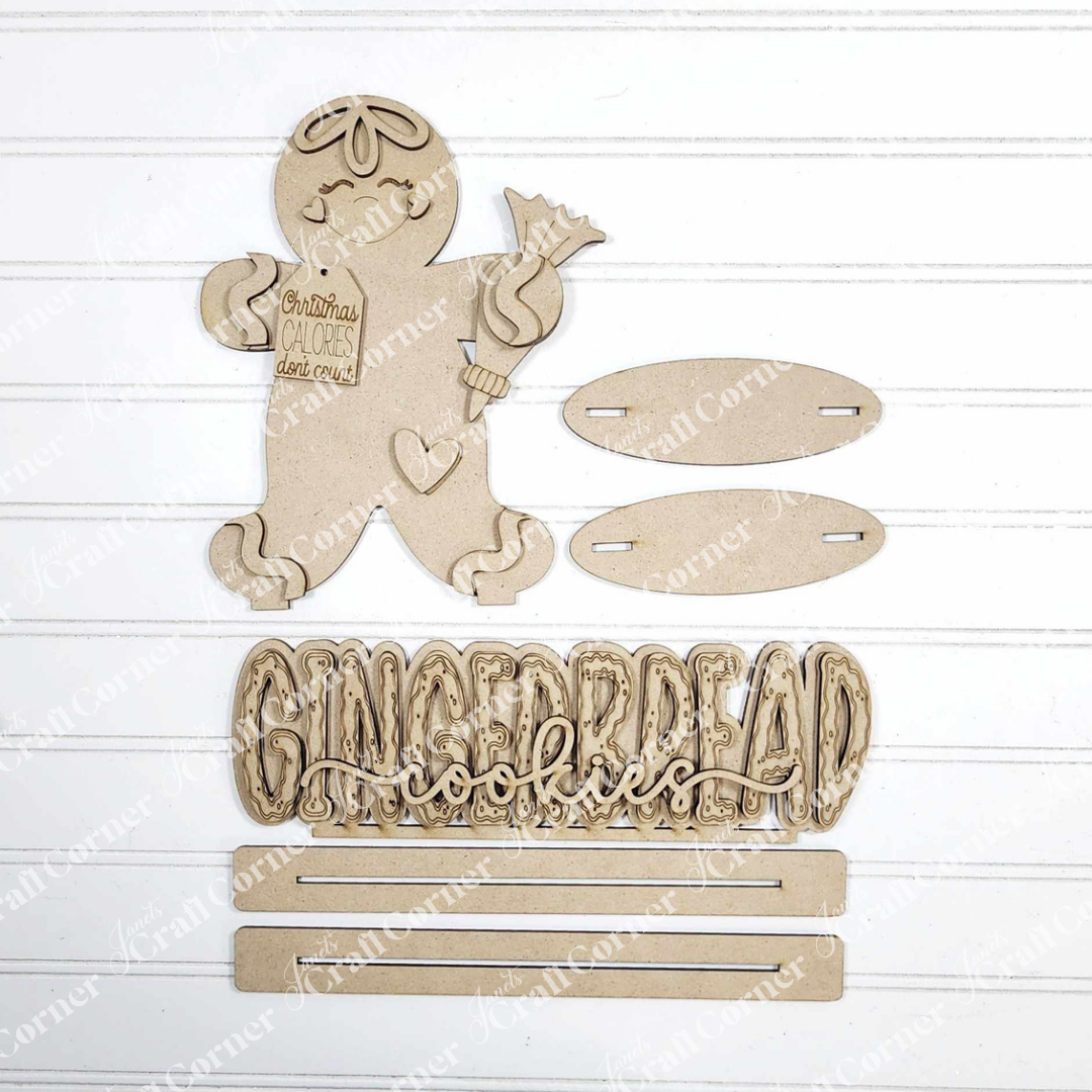 Janet's Craft Corner Gingerbread Baking Shelf Sitters DIY kit includes 2 unpainted wooden figures, a "Gingerbread Cookies" sign, slotted oval bases, and three strips—perfect for creating festive holiday home décor.
