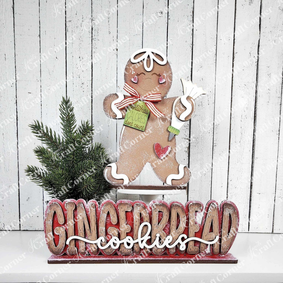 Janet's Craft Corner's Gingerbread Baking Shelf Sitters DIY kit (set of 2) features gingerbread figures with a rolling pin and recipe card beside a pine branch, “GINGERBREAD cookies” in faux snow on white wood—ideal for holiday décor.
