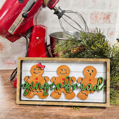 A wooden sign featuring three gingerbread figures, each decorated with red buttons and one wearing a red bow tie, showcases the word "gingerbread" in green script. A red mixer and pine branches provide a festive backdrop. This delightful piece is ideal for crafting enthusiasts interested in the Gingerbread Chunky Shelf Sitter DIY Home Decor Kit from Janet's Craft Corner.