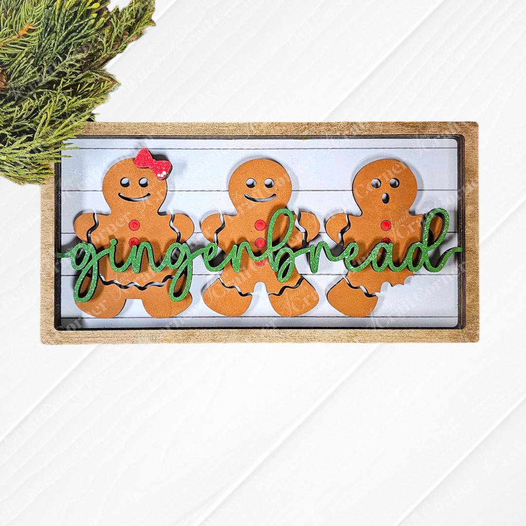 A wooden sign featuring three gingerbread figures, each decorated with red buttons and one wearing a red bow tie, showcases the word "gingerbread" in green script. Pine branches provide a festive backdrop. This delightful piece is ideal for crafting enthusiasts interested in the Gingerbread Chunky Shelf Sitter DIY Home Decor Kit from Janet's Craft Corner.