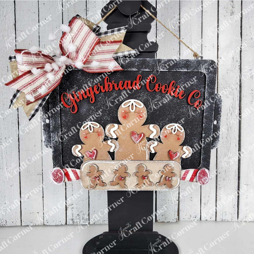The Gingerbread Cookie Sheet Sign by Janet's Craft Corner is a DIY seasonal home decor craft kit featuring "Gingerbread Cookie Co." in red, three smiling gingerbread people, small cookies, and a festive bow—perfect for holiday decorating.