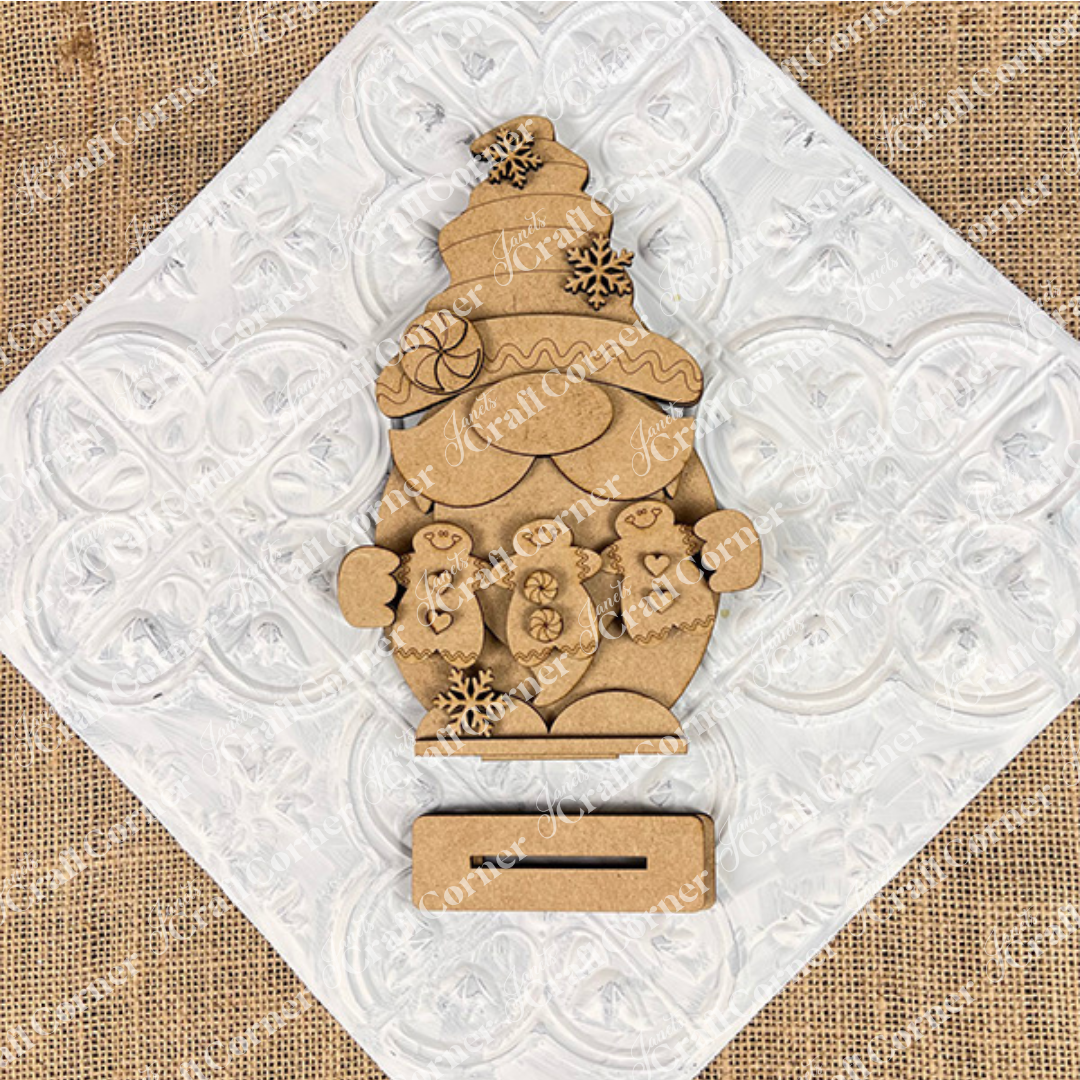 The Gingerbread Gnome Shelf Sitter by Janet's Craft Corner, featuring a festive hat and holding two gingerbread people, is adorned with snowflakes. Set against a white heart-patterned tile on burlap, this DIY seasonal decor craft kit adds charm to any festive setting.