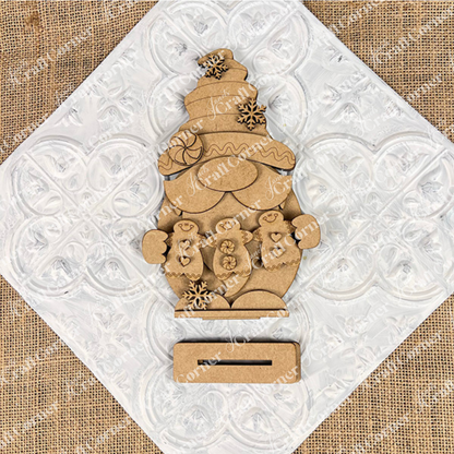 The Gingerbread Gnome Shelf Sitter by Janet's Craft Corner, featuring a festive hat and holding two gingerbread people, is adorned with snowflakes. Set against a white heart-patterned tile on burlap, this DIY seasonal decor craft kit adds charm to any festive setting.